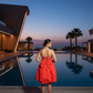 Woman in a red dress standing by a pool at dusk with modern architecture and palm trees in the background.