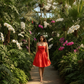 Woman in a red dress walking through a lush greenhouse filled with orchids.